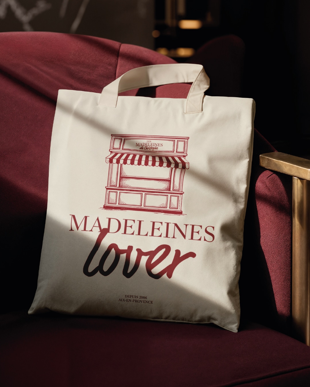 Sac tote bag en coton « Madeleines Lover » avec illustration de boutique et logo artisanal, posé sur un fauteuil en velours.